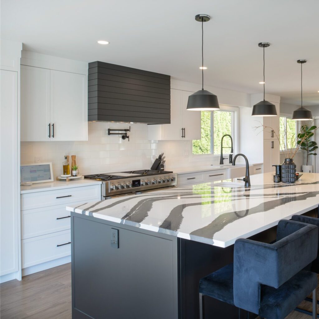 Kitchen view view with black and white quartz island countertop in foreground. Black fixtures like seating and an island sink can be seen. Three black accent lights hang over the island.