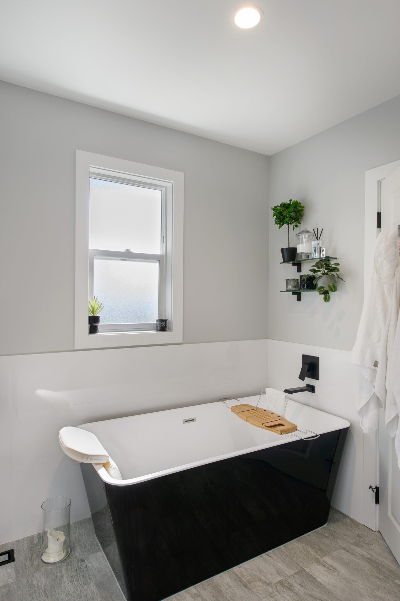 Freestanding with black exterior and white lining nestled into a corner by a rectangular single window, surrounded by a white ceramic tile backsplash and light grey walls.