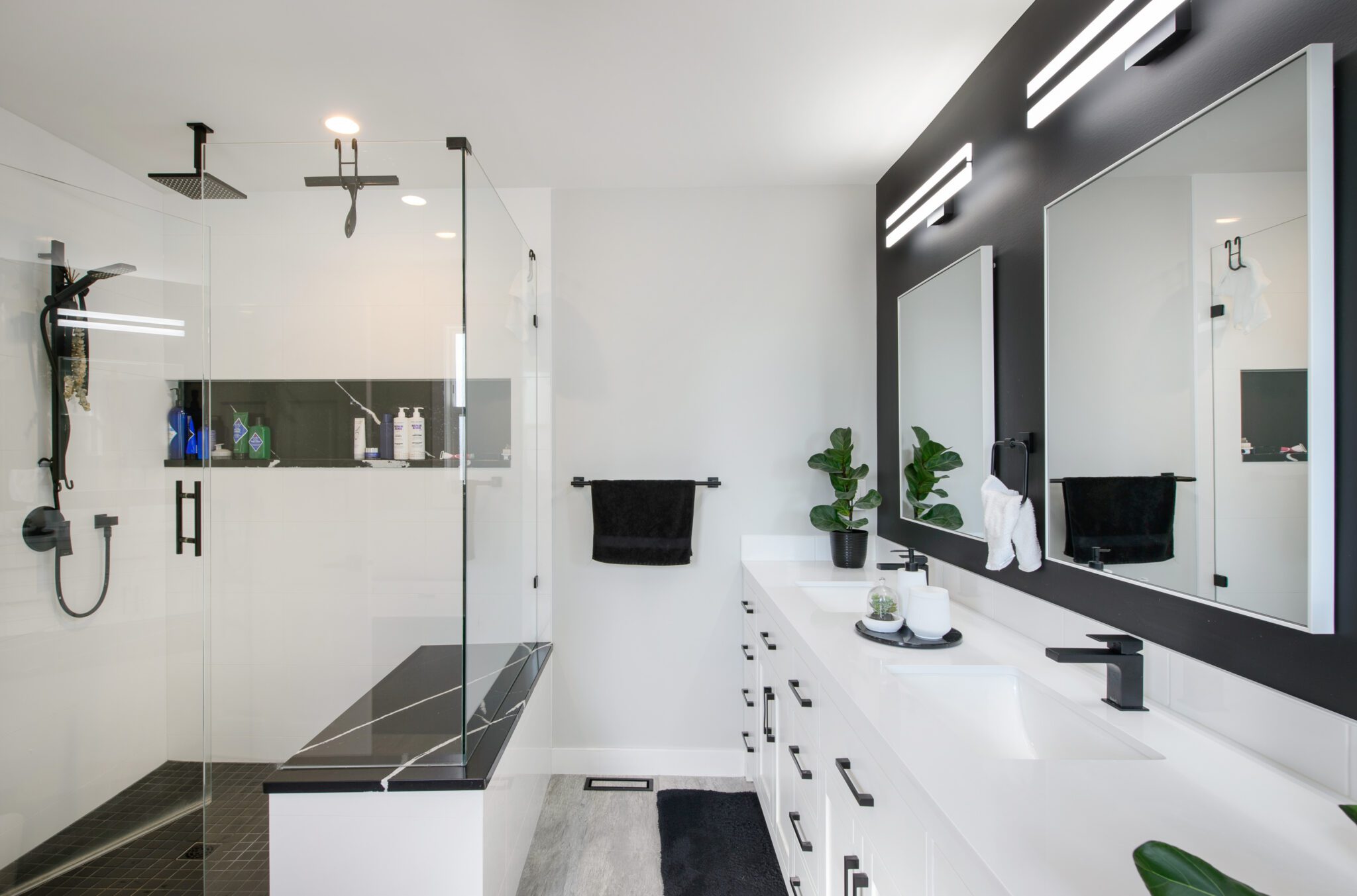 View of the bathroom shower with walk-in glass door and black, marbled shower seat. Across from it, a white vanity against a black wall.